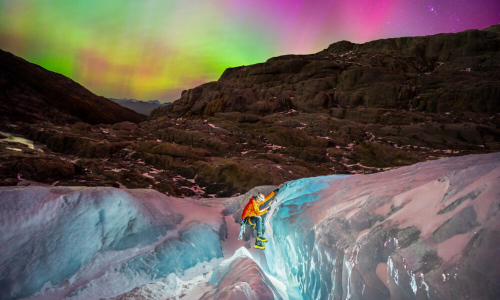 a glacier hiker climbs in the foreground with a headlamp as a rainbow colored aurora dances above